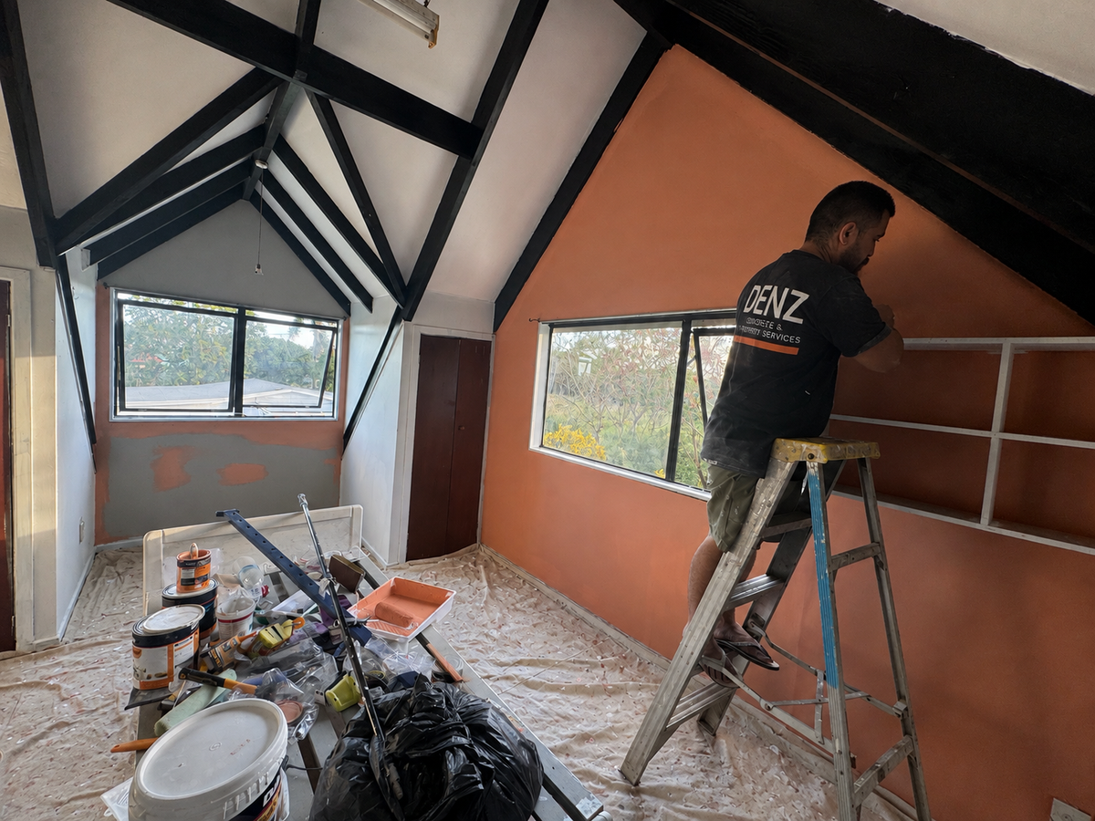 A man wearing a black t-shirt with 'DENZ' printed on the back climbs a metal ladder to install white wall shelves in a room under a steep gabled ceiling with exposed dark wood beams.