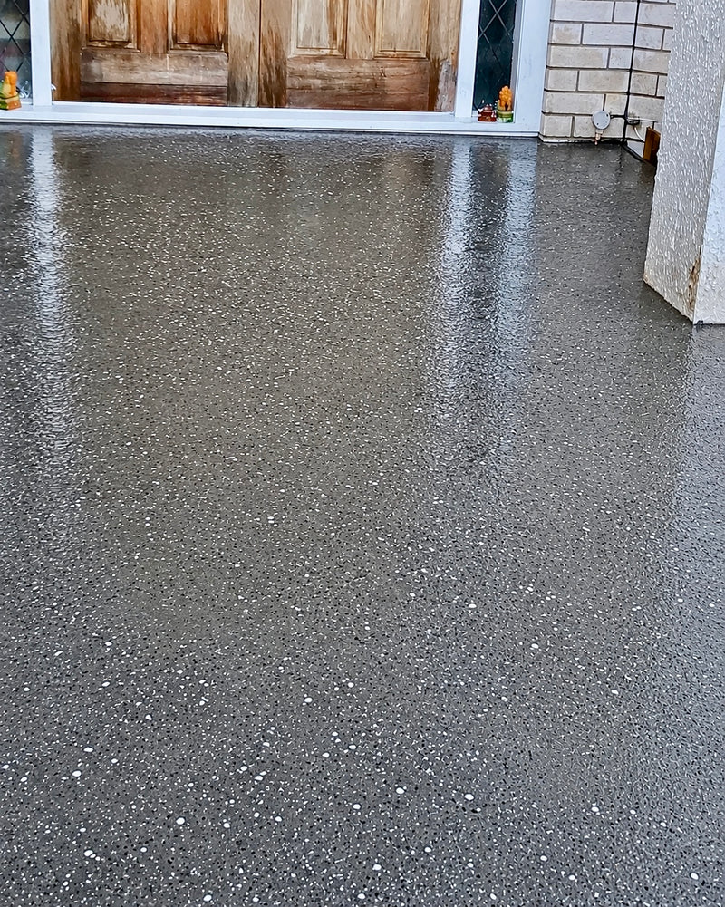 A wet gray exposed aggregate concrete driveway with small white and gray flecks, reflecting light near the top where a wooden door and light brick wall are visible in the background at a house in Melbourne 