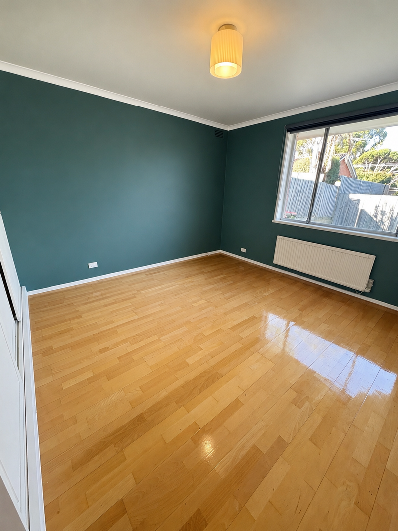 An empty room with glossy light brown hardwood floors and dark green painted walls, featuring a white ceiling, a cylindrical ceiling light, and a large window on the right wall.