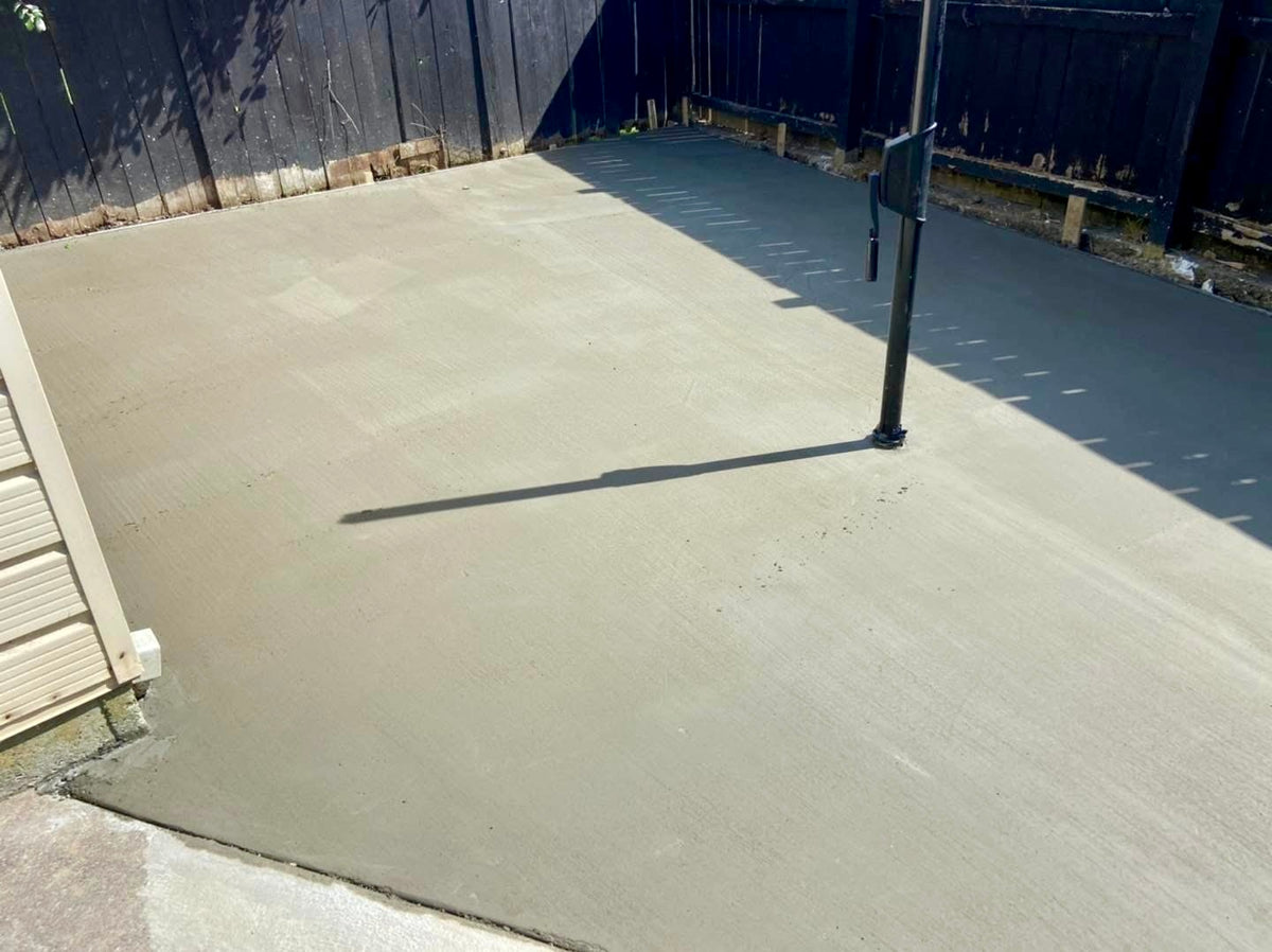 A freshly poured concrete patio surface in an outdoor backyard setting, with a dark wooden fence in the background and a black metal pole casting a long shadow across the concrete at a home in Melbourne.
