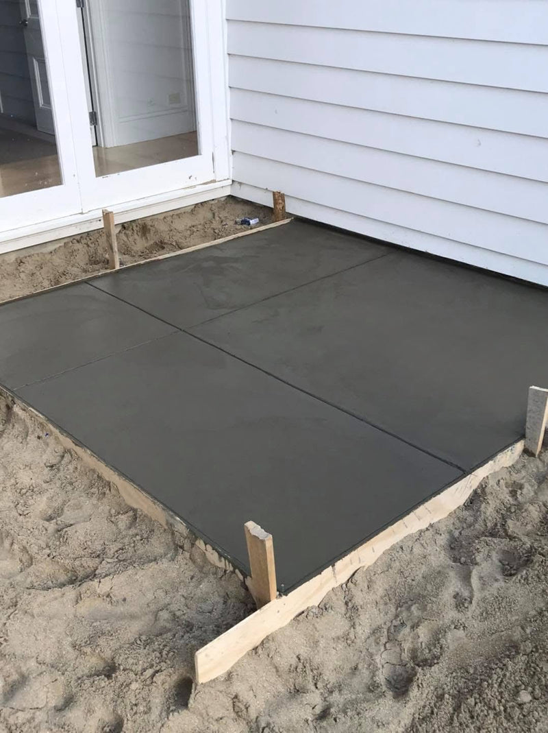 A freshly poured concrete patio slab in progress, shown outdoors next to a house with white siding and a glass door, with sand visible around the edges.