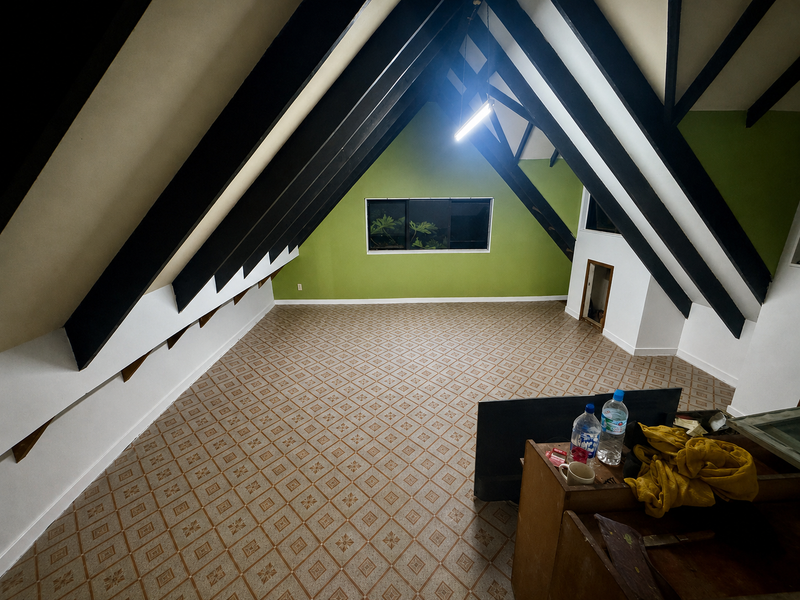 An empty attic room with a tiled floor, green accent wall, and prominent black wooden beams on a white ceiling. A small window is visible on the back wall, and a cluttered wooden table sits in the bottom right corner.