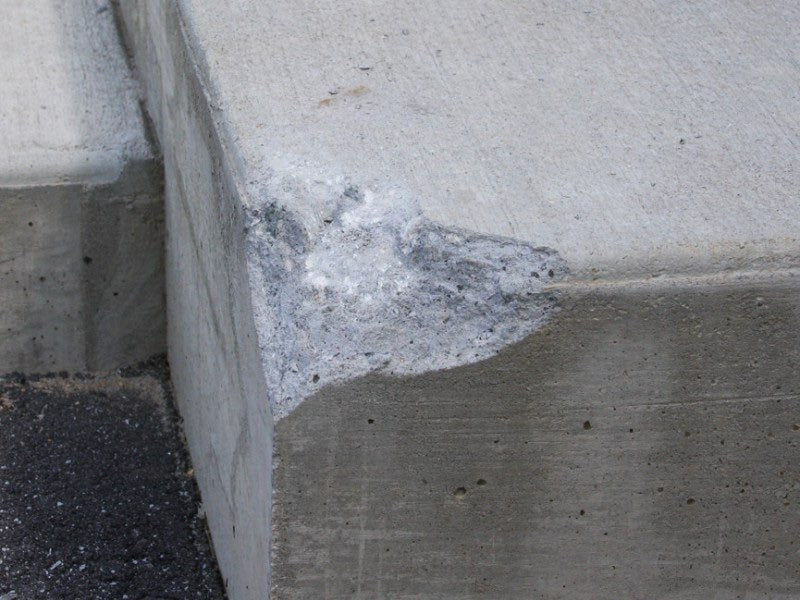 A close-up view of a concrete block corner with a rough, weathered patch of gray and white material near the joint, showing textured concrete surfaces and dark asphalt at the edge.