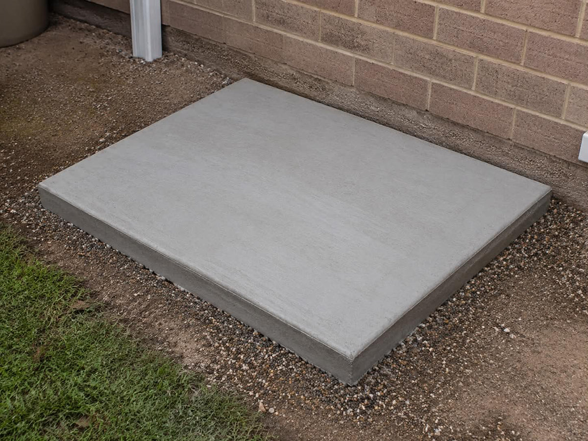 A square concrete slab foundation pad placed outdoors on a bed of gravel next to a brick wall and grass.