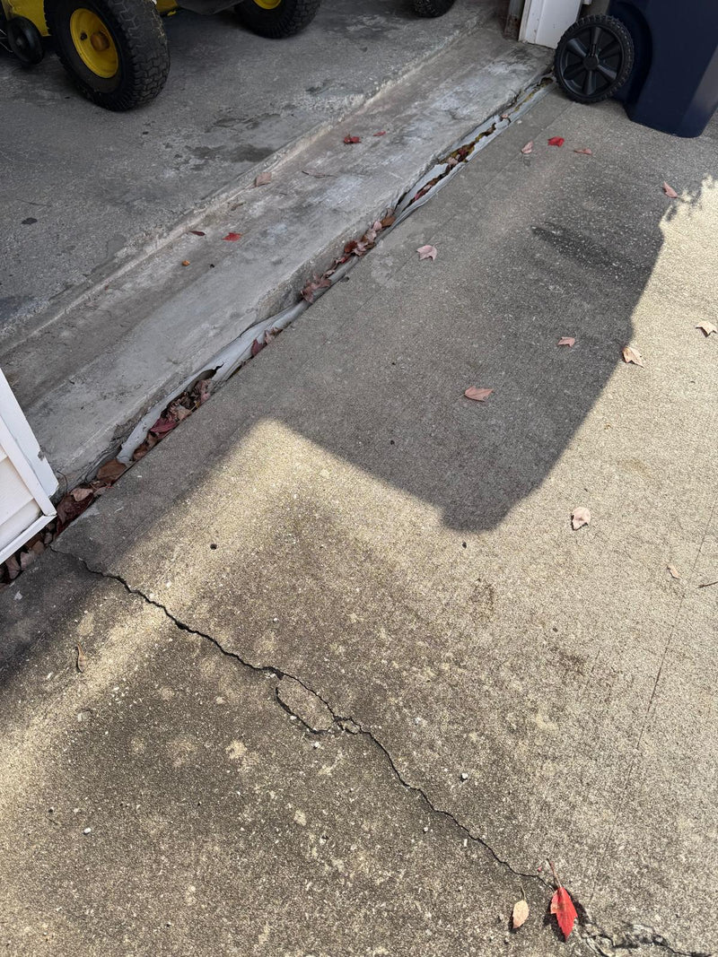 A close-up view of a cracked concrete driveway or sidewalk, showing a prominent fissure running diagonally across the gray pavement with scattered dry leaves.