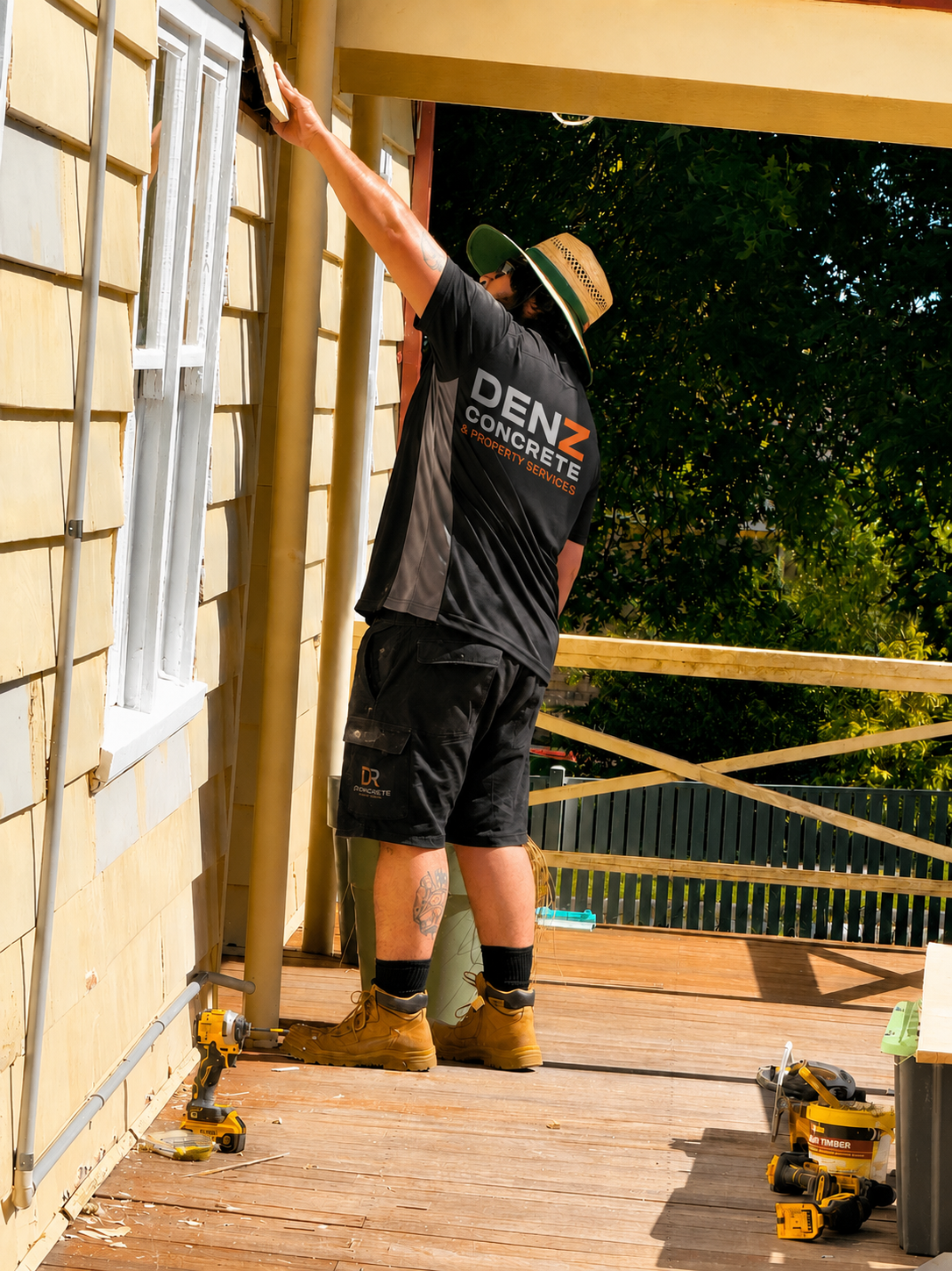 A construction worker wearing a black DENZ Concrete team shirt and shorts stands on a wooden deck, installing or repairing siding near a white window. Yellow power tools are visible on the deck, with trees in the background.