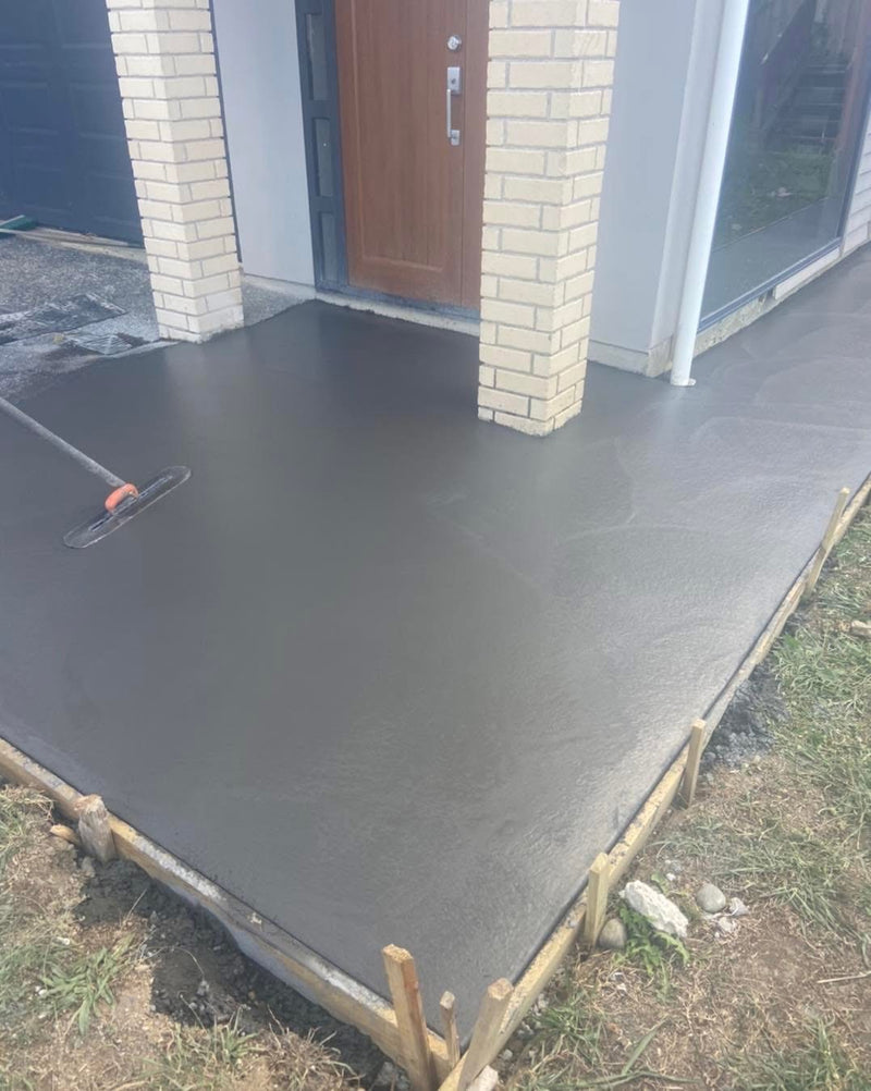 A freshly poured concrete porch floor with a wet finish, bordered by wooden stakes, with a house entrance, brick columns, and grass visible around the perimeter.
