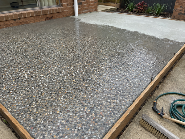A freshly poured concrete patio slab with embedded pebbles and stones, bordered by wooden edging, shown outdoors next to a brick house with a garden hose and brush visible on the ground.