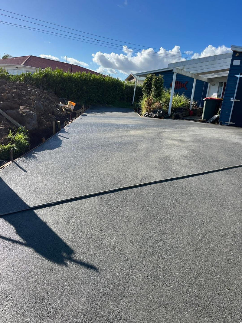 A wide concrete driveway or patio area in front of a small building with blue walls, white trim, and a hedge under a blue sky with clouds.