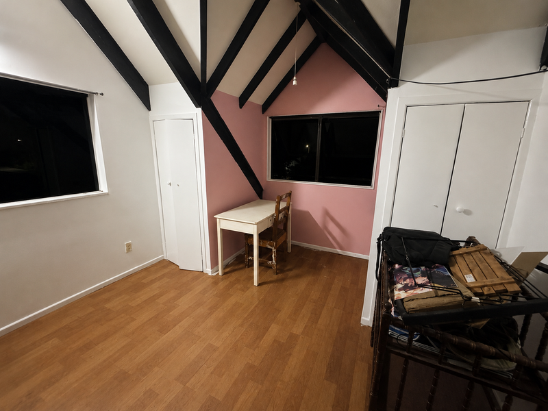 An attic room with a slanted ceiling, black exposed beams, light wood floors, a pink accent wall, a small white desk and chair, and white doors. A cluttered black storage cart is visible in the foreground on the right.