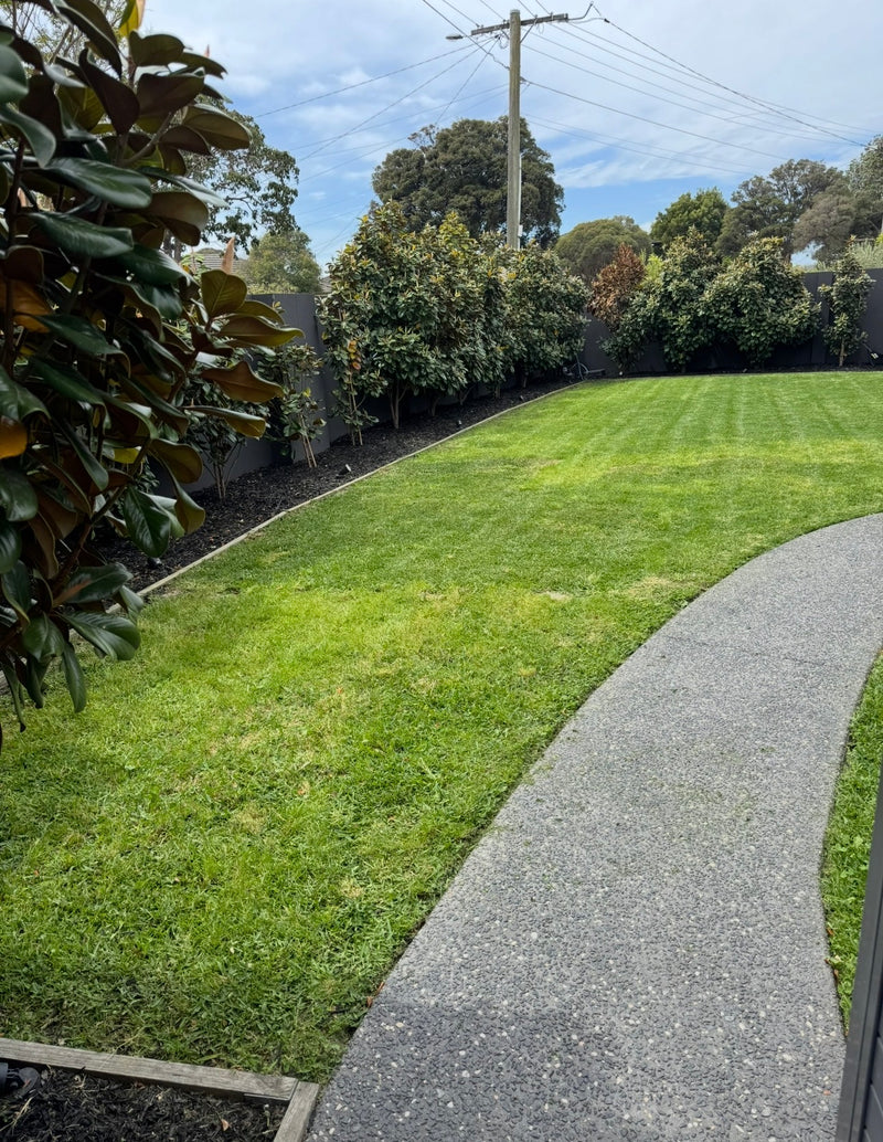 A well-maintained green lawn with a curved gray concrete pathway in the foreground, bordered by leafy shrubs and trees under a blue sky.