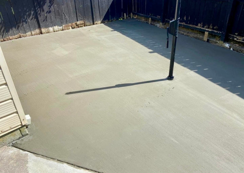 A freshly poured concrete patio surface in an outdoor backyard setting, with a dark wooden fence, a black metal pole casting a long shadow, and a beige siding edge visible on the left.
