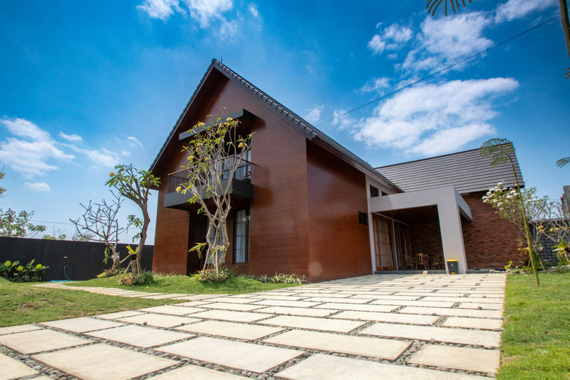 Modern single-story house with reddish-brown wood siding and a dark gray roof, set outdoors under a bright blue sky with white clouds. A stone patio leads up to the entrance, with grass, small trees, and a dark fence visible.