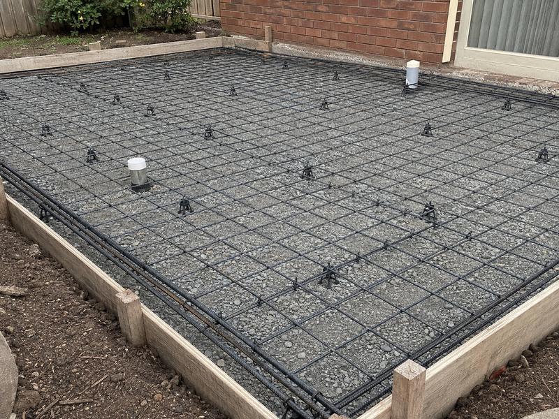 A rectangular concrete slab foundation with a black wire mesh reinforcement grid, filled with gray gravel. The slab is bordered by light-colored wooden forms and is set outdoors next to a brick wall and a glass door.