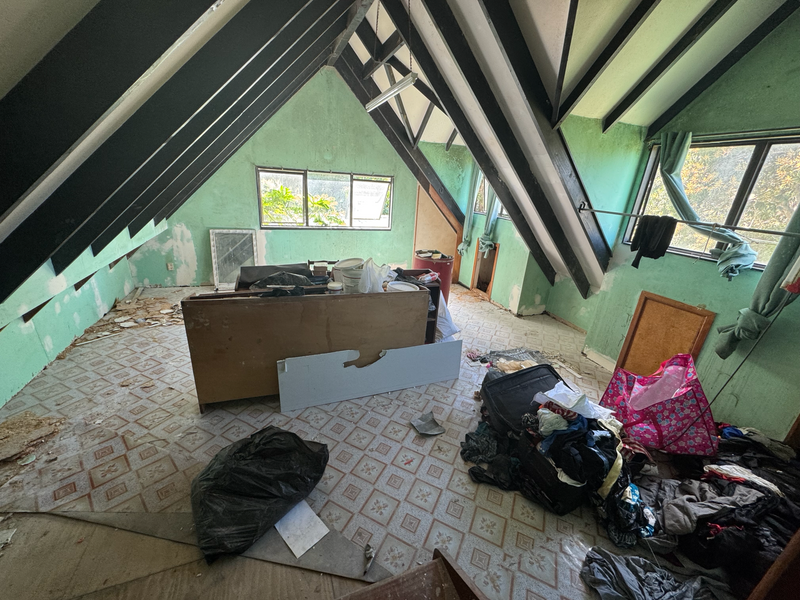 A cluttered attic room with green walls, exposed dark wood rafters, a patterned tile floor, a wooden desk or cabinet, and piles of trash, bags, and debris scattered around the space.