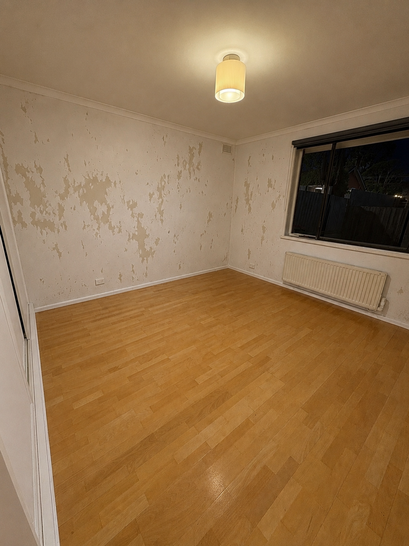 An empty room with light-colored wood laminate flooring, peeling beige wallpaper, a white ceiling light, and a window on the right wall.