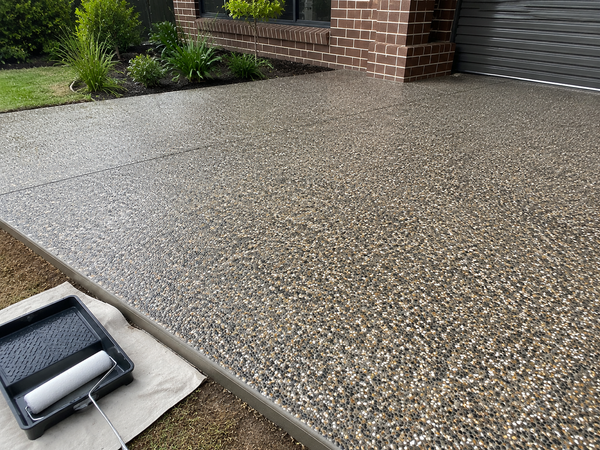 A freshly poured concrete patio with a textured aggregate finish, shown outdoors next to a brick house, garage door, and landscaping. A paint roller tray is visible in the lower-left corner.