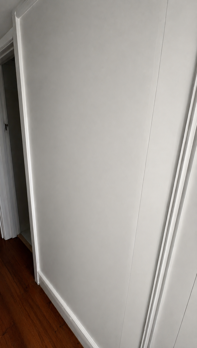 A close-up view of a white interior wall corner with white trim and baseboard, showing a dark wood floor in a doorway.